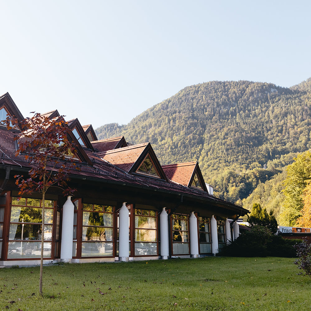 Außenansicht des Speisesaalbereichs: vor der wintergartenartigen Fensterfront mit weißen Säulen ist eine Wiese, ein Baum und Sträucher; Man sieht auch das Dach mit den Dreiecksgaupen und Blick in die grüne Berglandschaft.